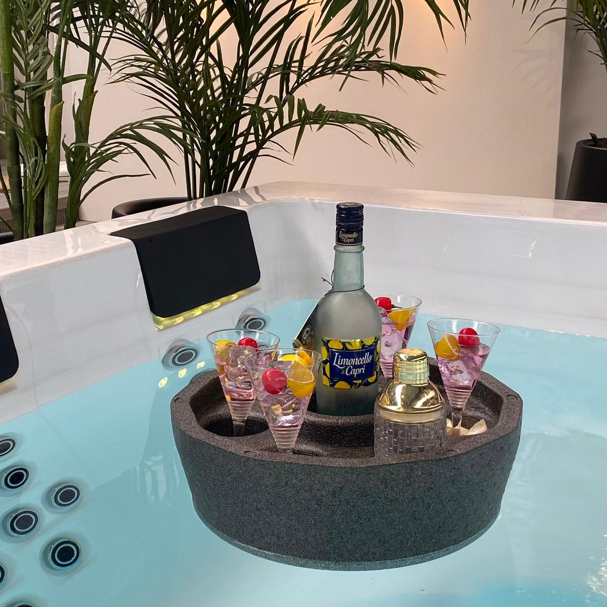 Floating bar with six cocktail glasses and bottles on dark foam tray, displayed in hot tub with tropical plants - Passion Spas Accessories